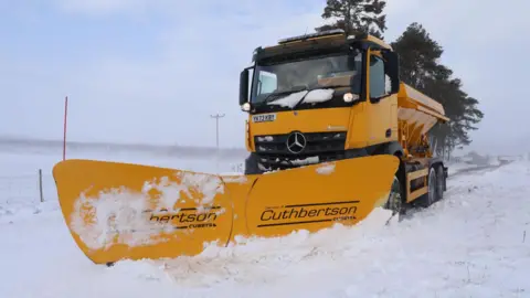 Peter Jolly/Northpix A yellow snow plough clears a rural road in Aberdeenshire. There are trees behind the vehicle.