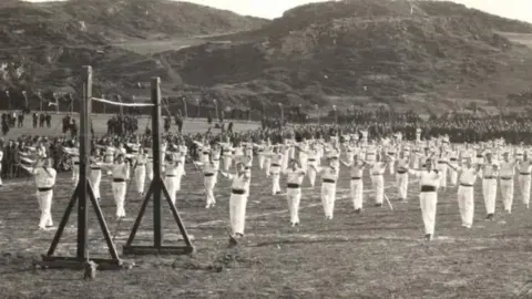 The mannin collection Hundreds of inmates, dressed in white, perform syncronised pilates. They're filed behind one another, all looking forward. The picture is black and white and was taken at the site of Knockaloe.