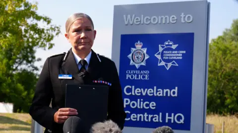 A police constable wearing a black suit, white shirt and black tie stands with a somber face, holding a clipboard, in front of a sign saying Cleveland Police Central HQ.