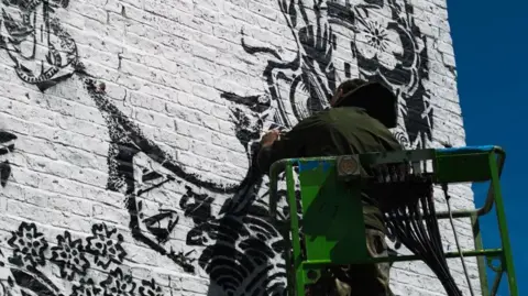 Close up of an artist painting a mural on the brick side wall of a house. He is seen from behind, standing on a railed platform, and wearing a dark green hooded jacket. A portion of the mural, in black on a white background, depits the lower part of a woman's profile surrounded by flowers. 