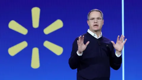 Getty Images Walmart Inc. President and CEO Doug McMillon in glasses and dark sweater holds up his hands while delivering a keynote address in front of the yellow Walmart logo during CES 2024 at The Venetian Resort Las Vegas on January 9, 2024 in Las Vegas, Nevada. 