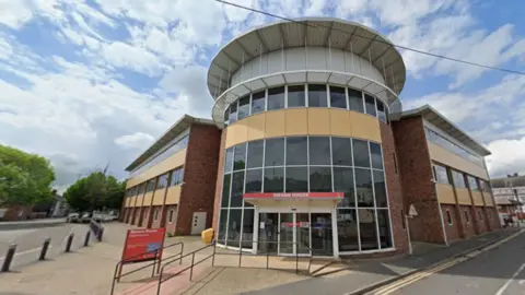 A three-storey building with a circuler glass atrium and red signage. The building is situated on a junction, and there are roads either side of it.