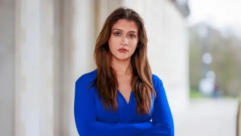 Liam McBurney/PA Wire Cara Hunter standing outside Stormont with her arms folded.   She has long, dark hair and is wearing a blue dress.  She is looking at the camera.