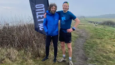 Two men in running clothes, one with a beard and curly hair and a blue coat and another in a blue t-shirt, cap and black shorts, stand on a moor next to a sign which reads 'Otley AC'