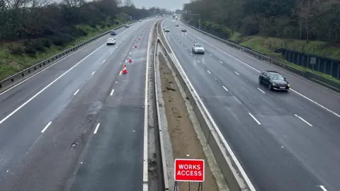 Tony Fisher/BBC A motorway with the outside lane southbound closed with cones. There is a sign in the foreground which reads: "Works Access". Trees and grass verges are on either side of the carriageway.