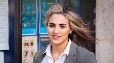 Dr Rahmeh Aladwan leaves court wearing a white shirt and light grey jacket. She has long fair hair and is wearing small gold hooped earrings