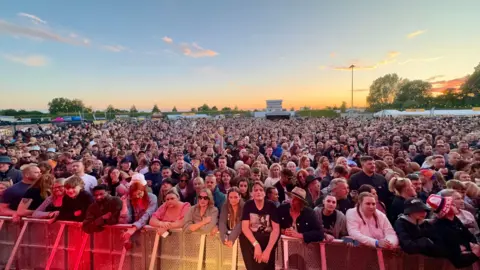 A sea of music fans watching the acts at this year's event as the sun sets. A red and yellow shadow from lighting can be seen in the foreground.