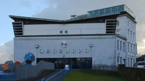 Galatas/Geograph A grey building with Catalyst written on the front of it. A fenced walkway leads up to the door, with blue and red shapes on the left.
