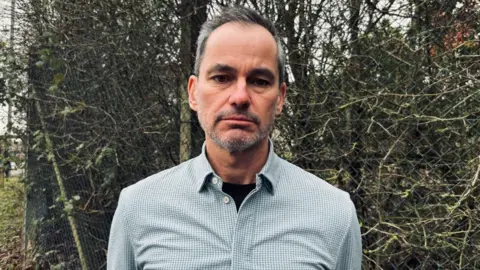 Getty Images Calvin Jones looks at the camera with a neutral expression. He is in a check shirt in front of a hedge