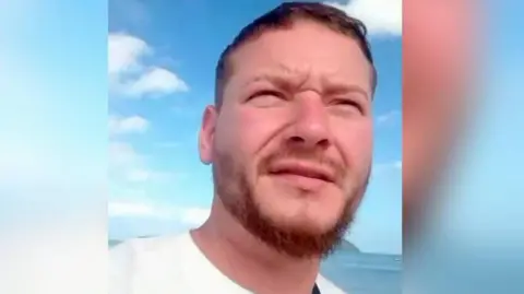 Mark Counsell, who has short brown hair and a beard and is squinting his eyes in the sun in a selfie, with sea and a mountain in the background. He is wearing a white top.