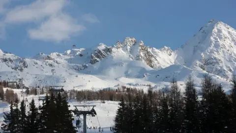 Getty Images Snow-covered mountains under a light blue sky. Tall trees and a ski lift can be seen in the foreground.