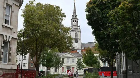 A street-level view of a Clerkenwell pedestrian square featuring a mix of historic stone architecture and lush green trees. In the centre, a white two-story building with a red door sits before a tall, multi-tiered church spire with a black clock face. People walk through the paved area, which is partially bordered by red wrought iron fencing and construction barriers under an overcast sky.