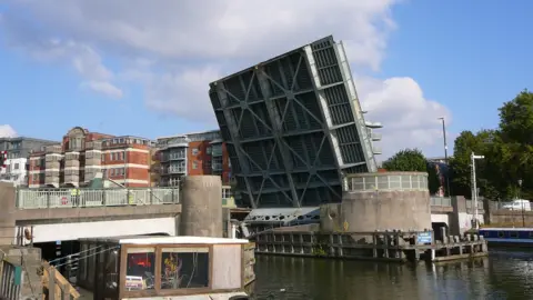 Bascule Bridge is lifted up to let a boat pass on the river underneath. In the background are red brick houses and trees.