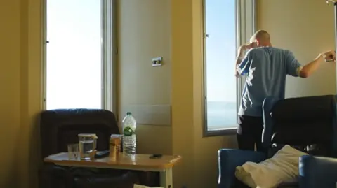 Éanna Mac Cana/Little Ease Films Éanna Mac Cana has his back turned to the camera as he stares out of the window of his hospital room. He is bald, and is wearing a light blue t-shirt and dark trousers. There are two armchairs in the room - one brown on the left and one dark blue on the right. There is a small table sitting in front of the left armchair with an empty bottle of water, a glass of water and a TV remote. Éanna has his arm up and is holding onto a metal pole and he brings his hand over his eyes.