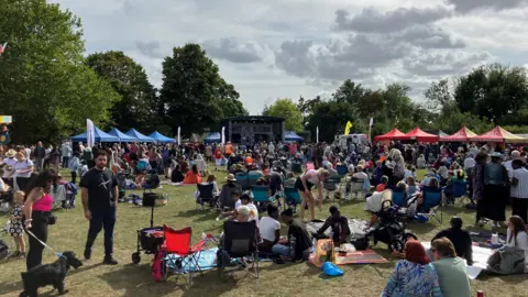 Jon Wright/BBC Crowds of people sit at an outdoor festivals with a mainstage in the background and different marquees around the edge