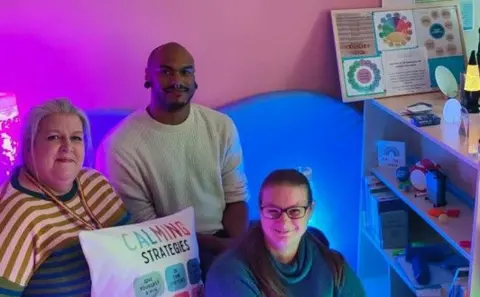 The picture shows a small sensory study space with calming features. There is a shelving unit holding items such as fidget toys, small containers, and books. The room is softly lit with coloured lights, including purple and green tones. There are three people in the room.