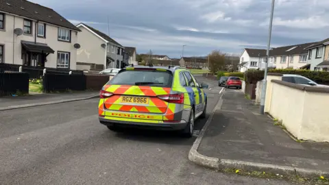 A police car sits beside a footpath in a residential area. The houses on either side of the road are two-storey, their facades painted off-white and their roofs covered in brown tiles.