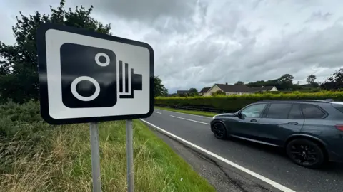 A speed camera sign on a grass verge next to a single carriageway road. A grey car is travelling along the road next to the sign. There is a hedge in front of a residential property on the other side of the road.