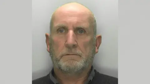 Gloucestershire Police A police mugshot of Michael Florek. He has a bald head with grey hair on the sides, and a grey beard. He is wearing a dark collared shirt and standing against a grey background with a blank expression.