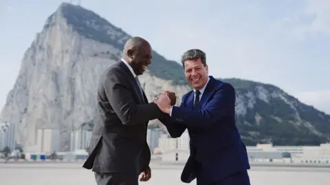 UK Foreign Secretary David Lammy and Chief Minister of Gibraltar Fabian Picardo clasp hands laughing, as they stand in front of the rock of Gibraltar.