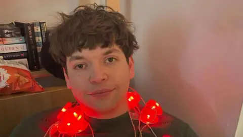 Handout Luis Piovesana wearing a dark grey jumper and sitting down in a corner, leaning up against a wooden bookcase. He has a string of red, love-heart shaped fairy lights around his neck. He has a dark brown, shaggy haircut and brown eyes, and is softly smiling at the camera.