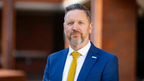Cleverbox Principal of Springwest Academy Simon Hart. He is wearing a blue suit and gold tie and smiling at the camera. He has a beard and dark, swept back hair.