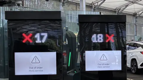 Two finger printing machines in the foreground of a hanger at Eurotunnel in Folkestone with cars and people in the background.