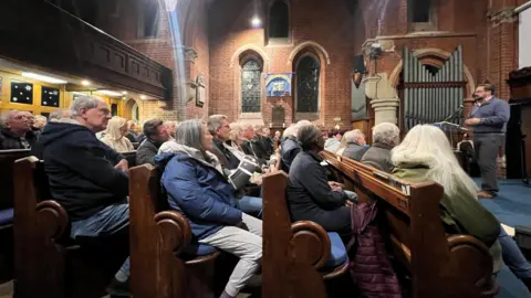 Carmelo Garcia Several dozen residents sit in church pews at a meeting sharing their concerns with councillors. One man is stood at the front, talking to them. He is standing near a set of organ pipes.