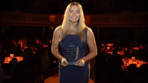 VANNIN PHOTOS Kaytlyn Adshead, who has long blond hair and it wearing an elegant blue dress, smiling. She is holding her trophy which is made of of glass and has a triangular peak. The tables at the Villa Marina can be seen in the background in low light.