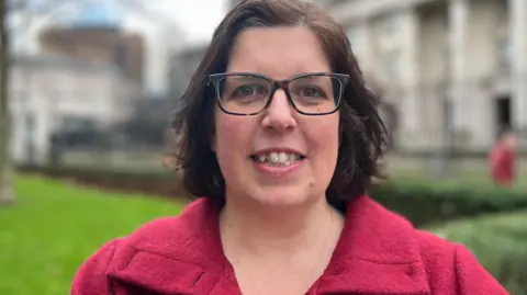 A woman looks into the camera smiling. She has short brown hair and is wearing a pink coat. She's standing outside.