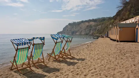 Getty Images Brightly coloured striped deckchairs on the beach on the Isle of Wight. The sand is golden brown and the sea is light blue. There are wooden huts on the beach and cliffs in the background. The sky is clear and it is sunny.