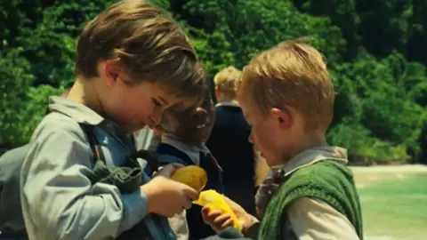 BBC/Eleven/Dualtagh Herr Two boys in school uniforms stand in a tropical clearing, examining and sharing a piece of yellow fruit while other boys gather behind them near the beach.