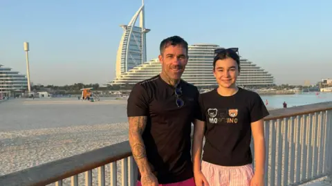 Ryan Hodgson stands next to his daughter in front of The Palm Dubai. They are both wearing black T-shirts. There is a beach behind them and blue skies. 