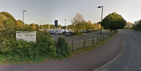 Google Sign which says Newmarket Road Park and Ride surrounded by bushes. A wooden fence is to the right and cars are parked behind it. 