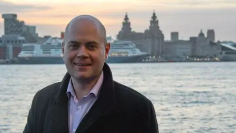 Liam Robinson, who is bald and is wearing a black woollen coat over a white shirt, smiles at the camera against a backdrop showing the Mersey estuary and the Liver buildings 