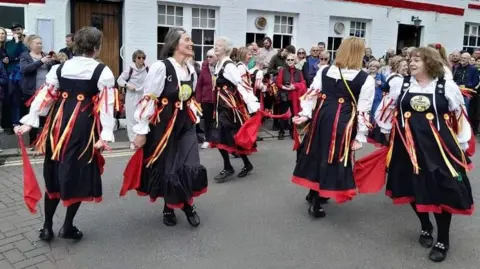 Sompting Village Morris Women wearing long black dresses, with red and yellow ribbons, spin and jump as part of a Morris dance to celebrate May Day. Spectators from the public watch on in the background.