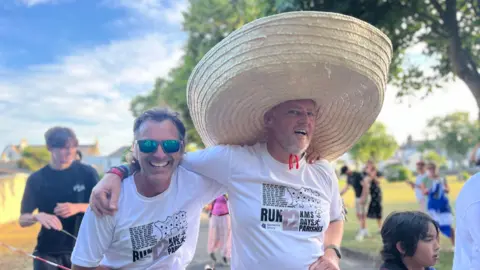 BBC Reverend Barrett crosses the finish line with an arm around another runner as they both smile and Mark is wearing a big sombrero