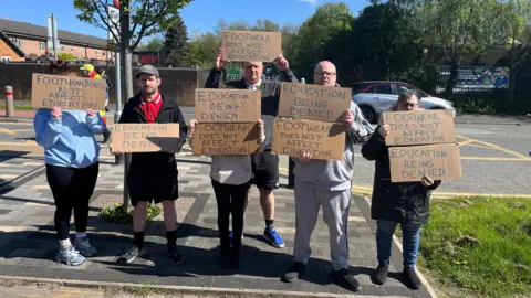 Several people stand around near the school holding handwritten cardboard placards saying 'Footwear does not affect eduction'