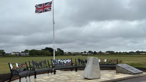 A view of the memorial stone, surrounded by seating with a union flag on a flagpole behind