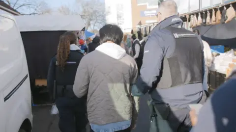 Home Office Two immigration enforcement officers escort a man through a market at Kempton Park racecourse in Surrey on 11 December.