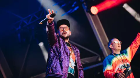 Nadine Ballantyne Goldie Lookin Chain performing at Bristol Sounds. Both men are wearing glasses and holding their arms up in the air.