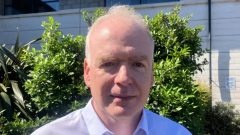 A man with white hair wearing a white shirt standing in front of a bush. The sun is shining.