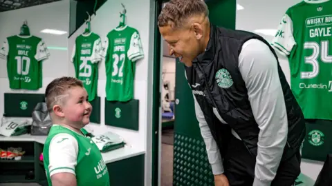 Sam Millar, eight, stands in the dressing room of Hiberian Football Club along with Dwight Gayle, one of the players. Sam is wearing a green football top and Dwight is wearing a black gilet jacket.