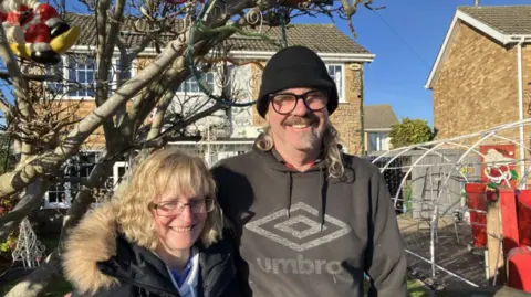 Lara King Couple standing in front of a house with Christmas decorations on the front of the house and in the garden. The woman who is wearing glasses has blonde hair and is smiling and has a blue coat on with a faux fur hood. The man is taller and has a black beanie hat on and is wearing black glasses and is smiling. He has long blonde/grey hair and a goatee beard with a moustache. There is a blue sky and they're standing next to a tree.