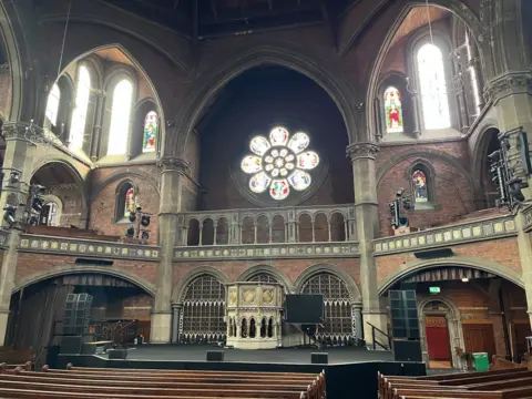 The inside of a large Victorian church with pupils leading up to a stage and pulpit with arched windows and balconies behind it