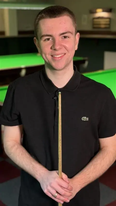 Oliver Sykes is grinning at the camera. He has short light brown hair. He is wearing a black polo shirt and holding a snooker cue, standing in front of a snooker table.