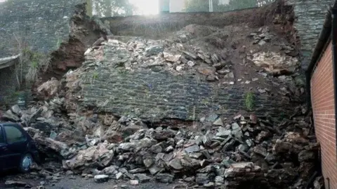 A large, tall section of a wall that has collapsed. There is a car in the bottom left of the image.