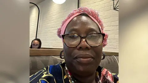 Ruth Mojisola Obasa A selfie of a woman wearing black glasses, a patterned top and a pink headdress. There's a plain white brick wall behind her