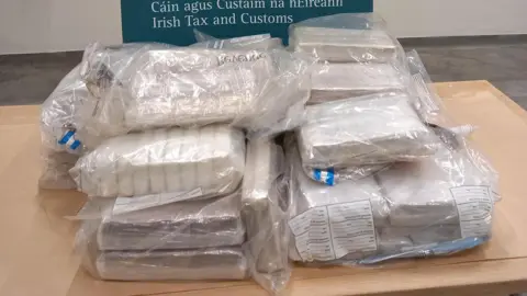 On top of a light-brown table sits dozens of white packages concealed inside clear colourless bags. A green banner reads "Irish Tax and Customs" behind the table.