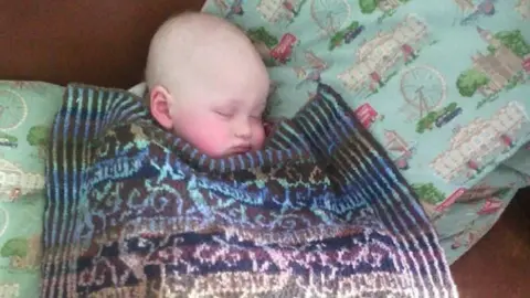 Gary Ray Smith A small sleeping baby tucked under a wool blanket which is multi-coloured. 